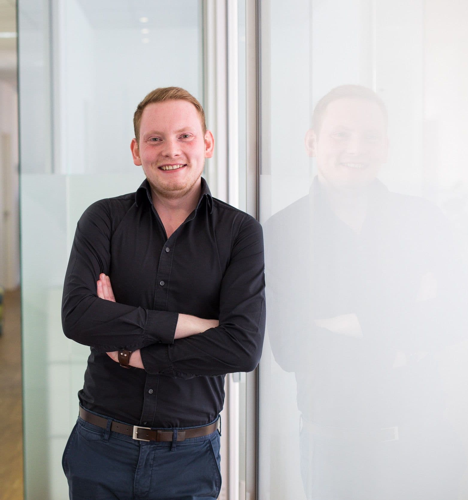 Maximilian Eckel, lächelnd an eine Glaswand gelehnt. Er trägt ein schwarzes Hemd und eine dunkelblaue Chino mit braunem Gürtel. 