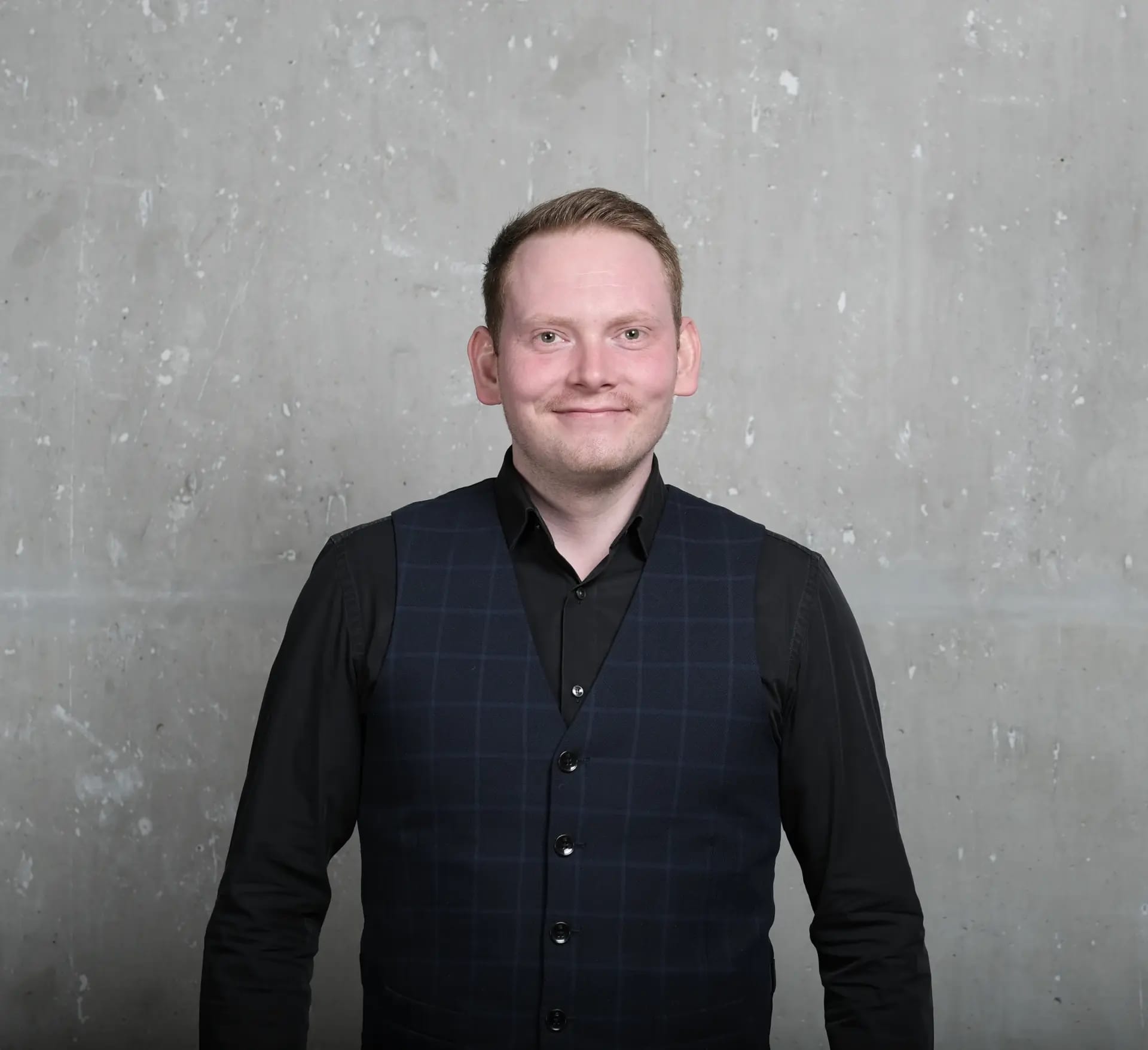 Maximilian Eckel, Gründer und Geschäftsführer von BespokeCode, vor einer grauen Betonwand. Er trägt ein schwarzes Hemd mit einer blau karierten Weste.