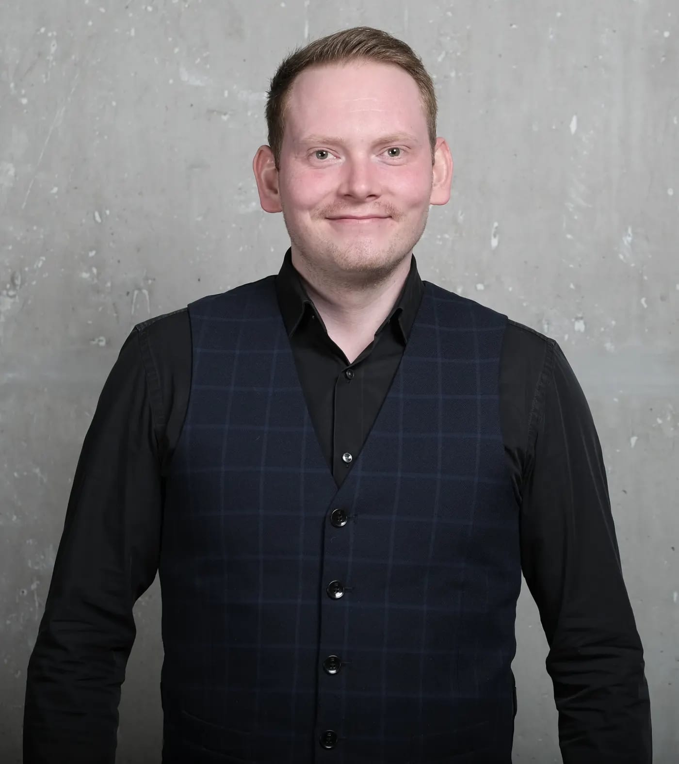Maximilian Eckel, lächelnd, er trägt ein schwarzes Hemd mit einer Blau-karierten Weste, vor einer grauen Betonwand